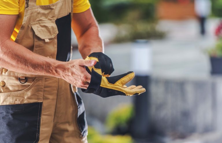 A construction worker puts on safety gloves, preparing for a task in a vibrant outdoor work area filled with greenery and tools. The worker is focused and diligent.